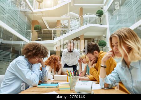 Two young optimistic girls cheerfully laugh at business meeting, keep hands on faces, hide eyes from strick boss, try to calm down, work time concept Stock Photo