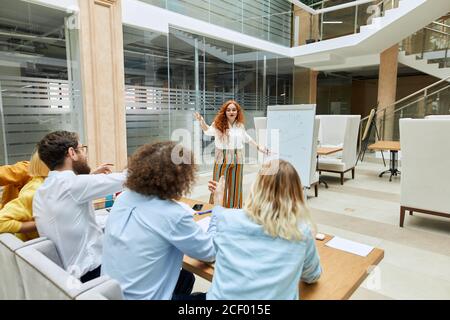 Pensive pretty girl wears white blouse, pointing at flip chart, stretches palm towards people waiting for answer, looks down thoughtfully, stands in t Stock Photo