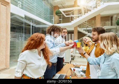 Team of young creative designers celebrate successful finish of job, stand around working table, cheer with happy expression, cooperative consept Stock Photo