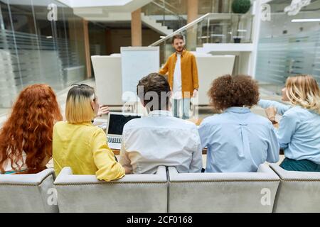 Photo of people sitting in soft comfortable armchairs with backs towards camera, listening to charming speaker attentively, team work concept Stock Photo