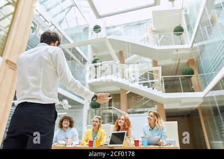 Back of tall brave man, formally dressed with nice black watch on hand, kindly talks to satisfied designers, spreads hand aside explaining strategic p Stock Photo