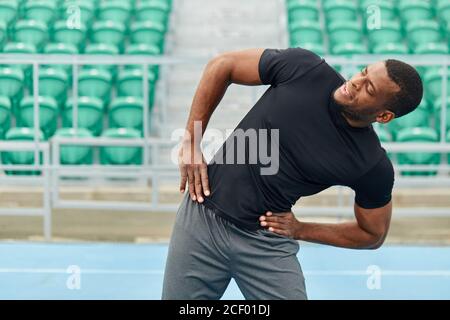african cheerful sportsman holding arms on the hips doing side bends outdoors. close up photo, colorful seats in the background of the photo. fitness, Stock Photo