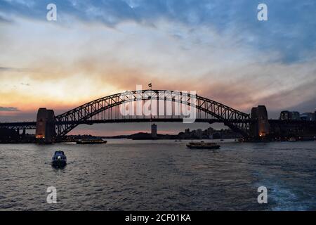 Sydney Harbour Bridge at sunset viewed from on the water of Sydney harbour.  Behind the bridge, Blues Point Tower and Blues Point can be seen. Stock Photo