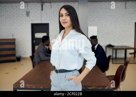 Caucasian beautiful woman wearing white formal shirt stand near table in office, confidently looking at camera