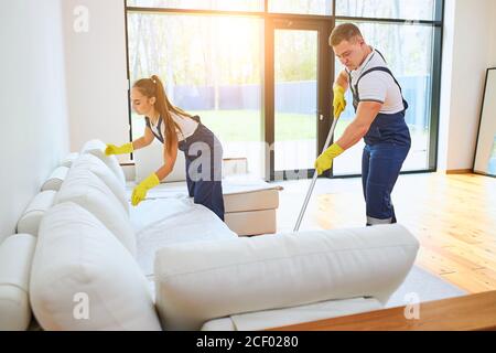 Two janitors in uniform cleaning new room with white sofa, mopping floor, straighten sofa. Work in country house, cottage Stock Photo