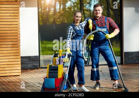 Two caucasian janitors, cleaners stand posing after successful job in big wooden house with panoramic windows. Holding mop, detergents Stock Photo