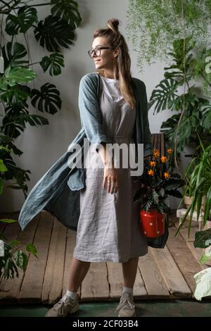 Happy woman gardener in eyeglasses wearing linen dress, holding a flowering calathea plant in old red milk can looking at window, standing in her home Stock Photo