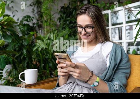 Close up of smiling positive woman sitting on chair in green house, resting, browsing surfing wireless internet on smartphone and sending message her Stock Photo