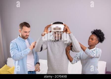 African young man trying on vr glasses for the first time and smiling diverse friends show him the right way to manage it indoor Stock Photo