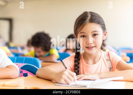 Portrait of cute little schoolgirl writing in book with classmates in ...