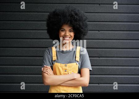 Smiling African hipster lady standing arms crossed against black wall. Stock Photo