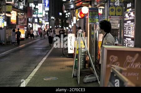 A server wearing a mask and face shield stands in front of a shop on a busy street near Kichijoji Station in Tokyo, Japan Stock Photo