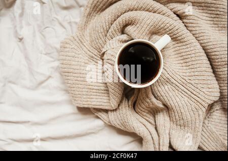 black coffee in white mug in knitted beige sweater on rumpled bed, top view, monochrome cozy picture of Breakfast Stock Photo