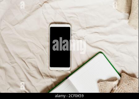 white smartphone with blank black screen, Notepad with white lined sheets and the sleeves of knitted sweater on beige crumpled fabric Stock Photo