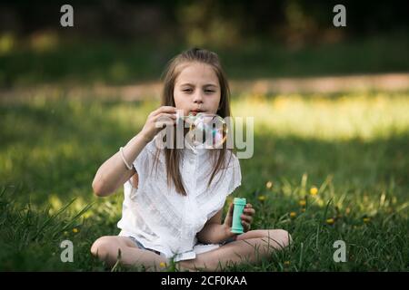 Little girl blowing bubbles sitting on the grass in the park. Stock Photo
