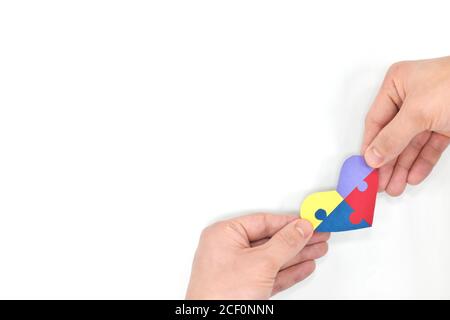 Male hands holding a colorful puzzle heart shape paper cutout. Top view flat lay in white background with copy space. World Autism awareness day, support and care concept. Stock Photo