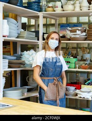 Waist up portrait of cheerful female artisan with face mask posing in pottery studio standing with arms crossed. New normal with social distance for small business and entrepreneur. Vertical Stock Photo