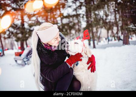 Young girl hugs dog with decorative deer horns Christmas labrador retriever in winter forest Stock Photo