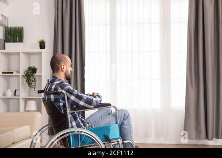 Handicapped freelancer in living room in wheelchair working on laptop . Serious sad caucasian man wearing casual clothes and look at large panoramic view in bright modern living room, hospital or clinic. Stock Photo
