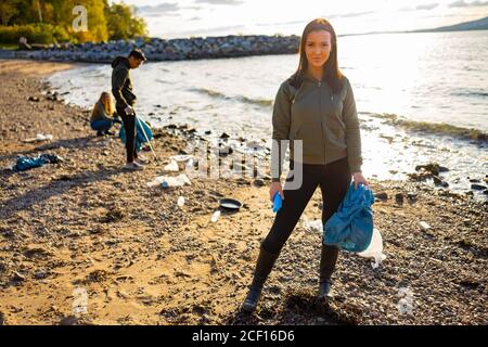 Smiling young woman cleaning beach with volunteers during sunset Stock Photo