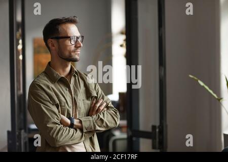 Young man in eyeglasses standing at office and looking through the window with pensive look Stock Photo