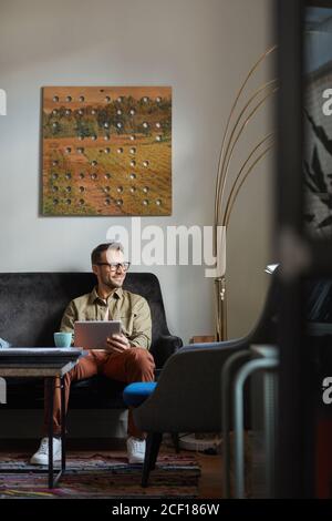 Young businessman in eyeglasses sitting on sofa and using digital tablet in his online work Stock Photo