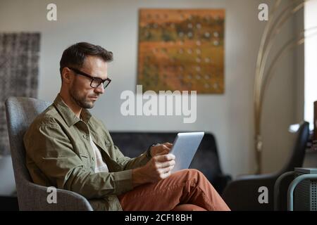 Serious designer in eyeglasses sitting on sofa and working online on digital tablet at the modern office Stock Photo