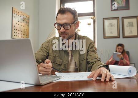 Mature businessman in eyeglasses sitting at his workplace in front of laptop and working over blueprint with his colleague in the background Stock Photo