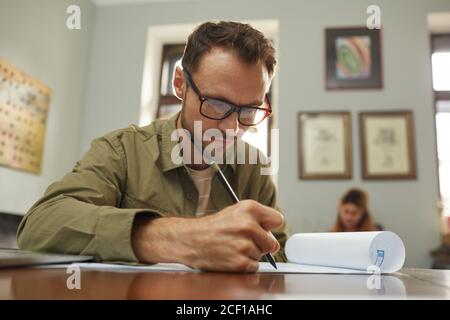 Mature designer in eyeglasses sitting at the table and drawing the blueprint of new project at office Stock Photo