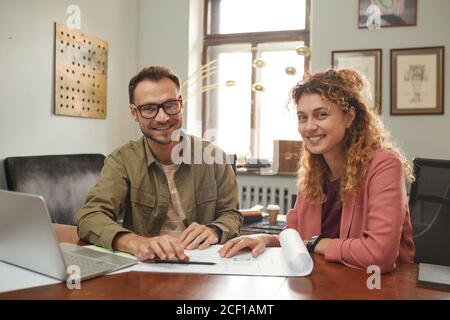 Portrait of two architects smiling at camera while sitting at the table with laptop and blueprint and creating the new project Stock Photo