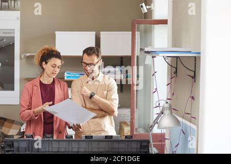 Young woman showing the blueprint in her hands and discussing it with her colleague while they working on printed machine Stock Photo