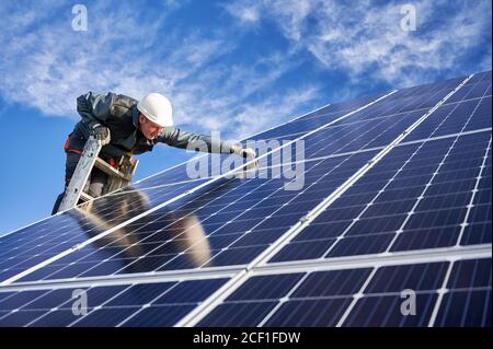 Male technician in white safety helmet standing on ladder and installing photovoltaic solar panel under beautiful blue sky. Concept of alternative sources of energy. Stock Photo