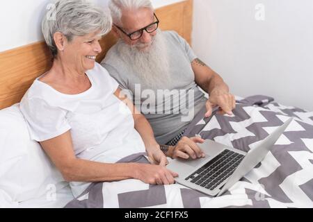 Happy married senior couple sitting in bed having video call on laptop - Mature people having fun at home - Technology elderly love and happiness conc Stock Photo