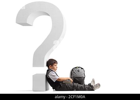 Sad schoolboy with a backpack and books seated on the floor and leaning on a question mark isolated on white background Stock Photo