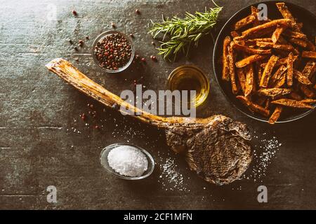 Grilled beef tomahawk steak with sweet potato fries salt spices olive oil and rosemary on rustic board - top of view Stock Photo