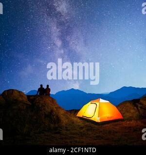 Young Couple Sitting on the Rock near Illuminated Tent and Watching the Bright Milky Way Above the Beautiful Mountains Stock Photo