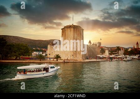 Touristic boat approaches Trogir harbor on the Dalmatian coast at sunset, with the tower of its medieval Venetian castle on the waterfront Stock Photo