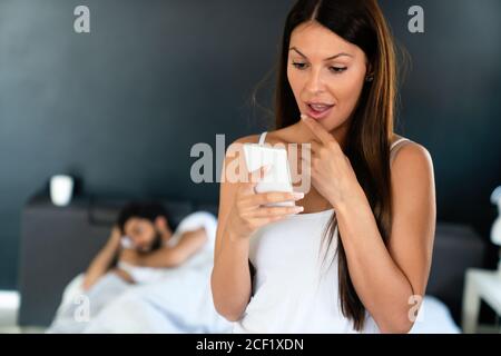 Young woman using cellphone while her husband sleeping on bed Stock Photo