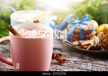 Christmas background of two mugs of hot chocolate with marshmallows, spruce branch and tray with gingerbread cookies on wooden table.