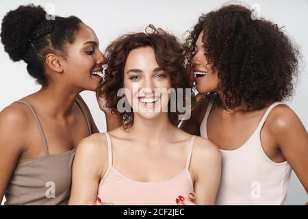 Portrait of three cheerful multiethnic women standing together and talking secrets in girl's ears isolated over white background Stock Photo