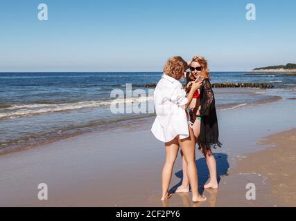 two girls in swimsuits take pictures of each other with smartphone on the seashore on sunny summer day Stock Photo