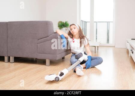 woman using vacuum cleaner in the living room.Young girl cleans the apartment. Tired girl after cleaning sits on the floor with a vacuum cleaner. Stock Photo