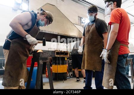 Busy blacksmiths in masks forging metal detail while hitting hot iron with hammer Stock Photo