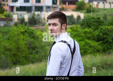 Portrait of stylish brunet young man, wearing white shirt, black tie and suspenders, walking in a park with lush foliage. Stock Photo