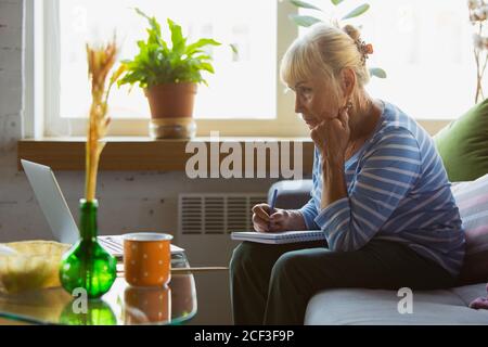 Making notes during lesson. Senior woman studying at home, getting online courses, self-development. Caucasian woman using modern devices for having fun, education, spending time for new job or hobby. Stock Photo