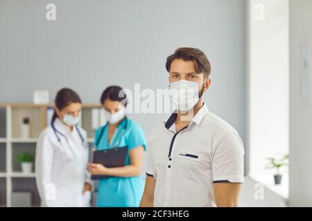 Man patient in medical protective mask standing over talking women doctors at background in clinic