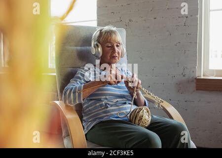 Learning to knit with an online lecturer. Senior woman studying at home, getting online courses, self-development. Caucasian woman using modern devices for education, spending time for new job or hobby. Stock Photo