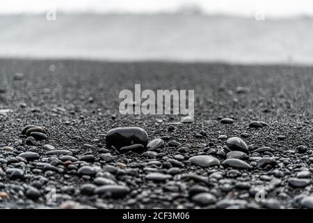 Closeup foreground of large black volcanic stones or pebbles on sand beach in Reynisfjara, Iceland with water waves crashing on shore to shiny wet roc Stock Photo