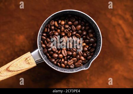 Top view of the cezve with a wooden handle in which are roasted natural coffee beans, and the cezve stands on the surface of ground coffee. Stock Photo