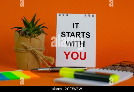 White note with inscription 'it starts with you' on beautiful orange background, colored paper, metalic pen, yellow marker,  flowerpot with a plant an Stock Photo
